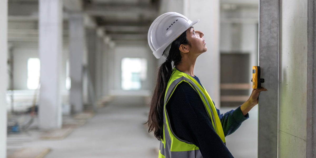 Female construction worker with safety vest and white hard hat measuring wall with special tool